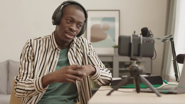Medium shot of young African-American male rap singer wearing earphones sitting at daw at home, rapping while recording it on smartphone camera