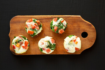 Homemade Egg White Breakfast Cups with Spinach and Tomato on a rustic wooden board on a black background, top view. Flat lay, overhead, from above.