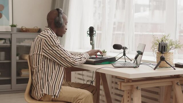 Medium shot of young African-American man wearing headphones sitting at m-audio keystation at home and making electronic music using laptop and synthesizer