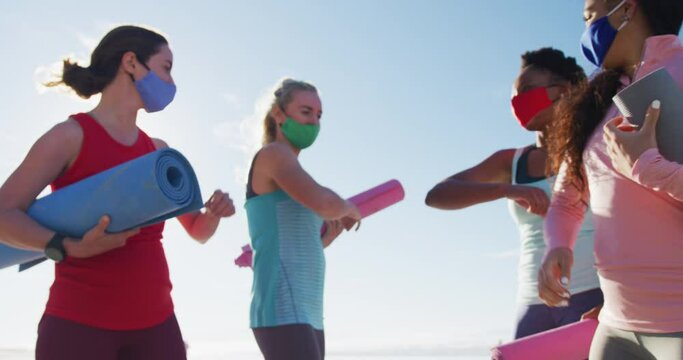 Group of diverse female friends wearing face masks holding yoga mats at the beach touching elbows