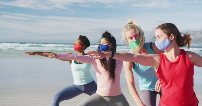 Group of diverse female friends wearing face masks practicing yoga at the beach