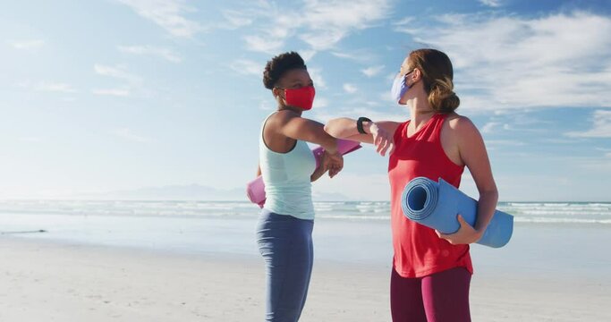 Two diverse female friends wearing face masks holding yoga mats at the beach touching elbows