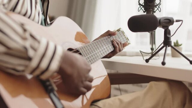 Close up of unrecognizable African-American man wearing striped shirt sitting in home studio in front of microphone and playing guitar