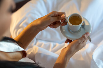 Fototapeta na wymiar A young female, wearing a skin-colored nightdress and holding a cup of coffee in her hands in bed on a white blanket. Concept of a relaxed morning in bed.
