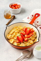 Mini white pancake cereal with strawberries in frying pan for breakfast on grey background . Trendy home breakfast with tiny pancakes. Top view