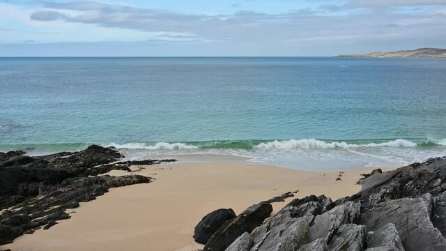 4k slow motion clip of wave crashing on pristine sandy beach with black rock either side of sand. Turquoise water, blue sky and cloud. Sunny day. Near Hargabost, Isle of Harris, Scotland.