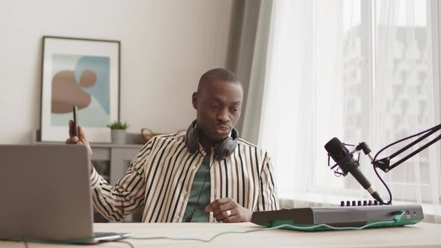 Lockdown of young male African-American artist sitting at modern midi keyboard synthesizer at home, playing on it while having video phone call