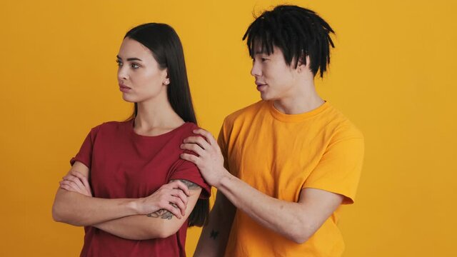 A multinational couple man and woman are standing isolated over a yellow wall in the studio. A displeased woman is offended, and a man is trying to calm down her