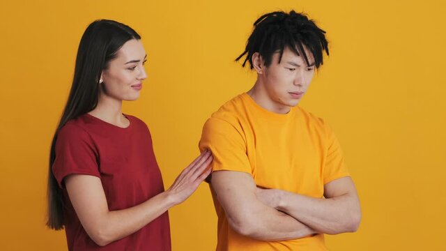 A multinational couple man and woman are standing isolated over a yellow wall in the studio. A displeased man is offended, and a woman is trying to calm down him