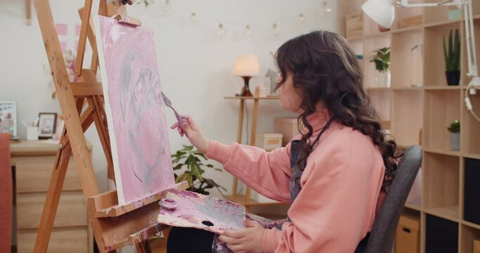 Side view of young creative girl making modern art picture using spatula and acrylic paints. Teenager artist with down syndrome sitting in front of molbert and creating .Indoors.