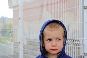 Portrait of a sad three-year-old boy in a hood against the background of a fence. The offended...