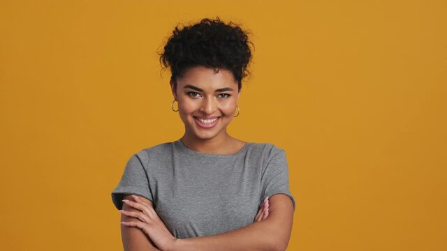 A positive american woman wearing gray basic T-shirt is posing to the camera standing isolated over yellow wall in the studio