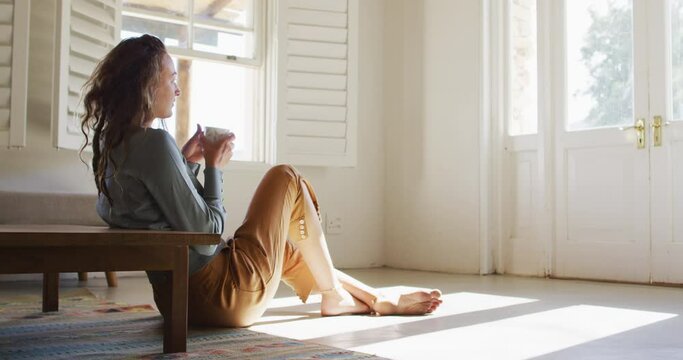 Thoughtful caucasian woman sitting on floor in sunny cottage drinking coffee, looking out of window