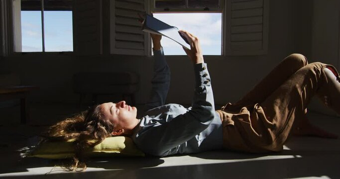 Caucasian woman lying on floor holding book and reading in sunny cottage living room