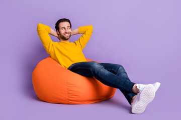 Full length photo of cheerful happy relaxed young man sit bean bag look empty space isolated on...