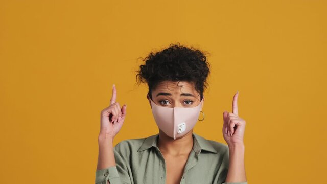 A close-up view of a confident american woman is wearing a facial medical protective mask pointing to the top standing isolated over yellow wall in the studio