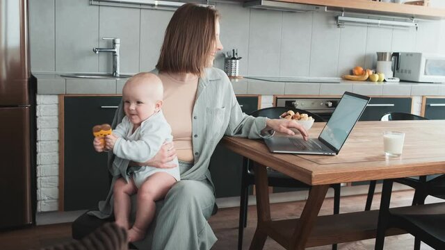 A positive mom sitting at a laptop and holding her little baby with a pacifier in her hands in the kitchen