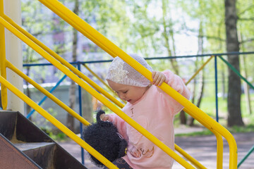 Stylish baby girl 2 years old in a pink fluffy jacket climbs the stairs to the children's slide on the playground