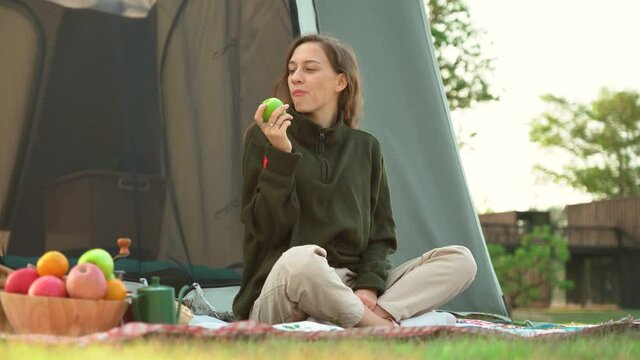 young hipster woman Sitting on mat in front of the tent and eating green apple to breakfast on countryside in nature park in morning outdoors in picnic camping
