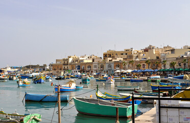 Fototapeta na wymiar Overlooking the harbor with traditional blue fishing boats and the ancient buildings of Marsaxlokk. Marsaxlokk is a small, traditional fishing village in the South Eastern Region of Malta.