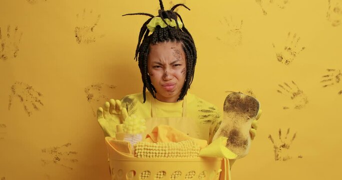 Discontent Afro American woman looks with aversion at dirty things smells stench frowns face holds dirty sponge poses near basket full of cleaning supplies brings house in order. Housekeeping