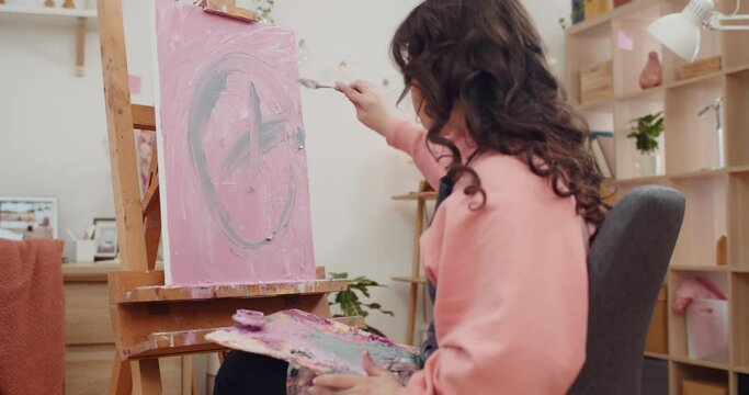 Side view of young female artist painting without brushes and oil. Teenager girl creating picture with palette knife while sitting in front of molbert in her room.Concept of creativity.