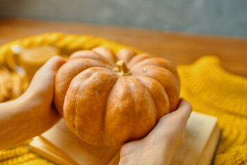 Autumn harvest festival. Girl holds a pumpkin over an open book, on a knitted sweater. Wooden table and concrete background.The concept of thanksgiving or Halloween.