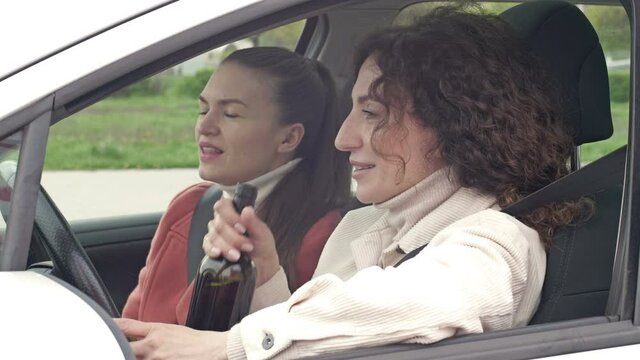 Two girlfriends drink wine from a bottle while sitting in the car. Drunk driving.