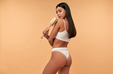 Young Asian woman in white lingerie makes anti-cellulite massage with a dry brush on a beige background. Body scrubbing, weight loss, body care, spa treatments.