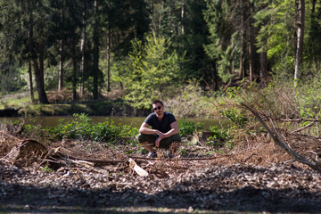 Attraktiver junger Mann hockt sich auf dem Boden im Wald. Er hat Sonnenbrille auf und genießt das Leben, Freiheit und schönes Wetter. Überall liegt Holz und die Äste. 