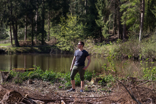 H&uuml;bscher Mann mit schwarzen Haare und Bart steht  sehr entspannt im Wald, hinter Ihm ist See. Er schaut zur Seite in die Ferne. Er hat Ziele und Pl&auml;ne, denkt nach, sieht motiviert und sportlich aus.