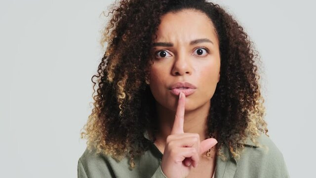 A shocked african american woman with afro curls is doing silence gesture isolated over a white wall in the studio
