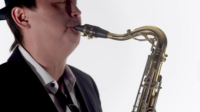 Man in a hat, jacket and tie blows a saxophone on white background in the studio, close-up. Male musician plays the saxophone.