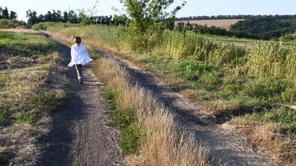 Defocused silhouette of little girl running away by dirt road at rural landscape in summer. White clothes fly in wind. Serenity of childhood