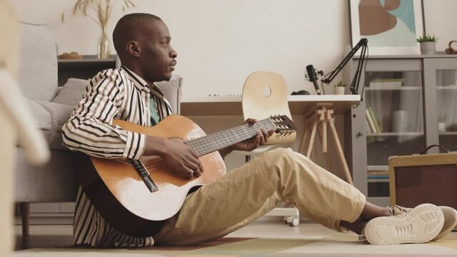 Wide shot of attractive African-American male musician sitting on floor in living room leaning on sofa near portable stereo speaker and enjoying playing acoustic guitar