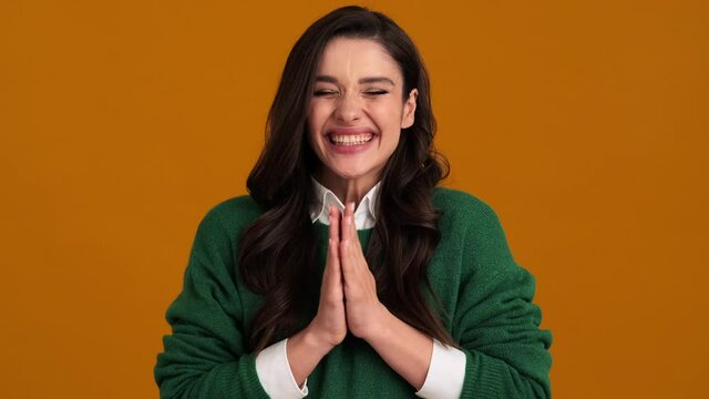 A happy woman in a green sweater and shirt bouncing and clapping while standing in an orange studio