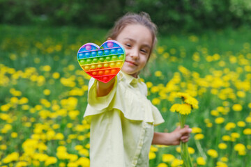 The girl holds in her hands a colorful pop it game in the shape of a heart. Popular pop toy in rainbow colors. Many dandelion flowers in the park.