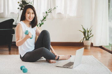 Healthy beautiful Asian woman sitting on the floor holding dumbbell using laptop at home in living room ready to online workout - Powered by Adobe