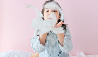 Closeup portrait of a cute little girl in pajama blowing white feathers while playing in the bed, on pink background. A kid playing games in the morning.