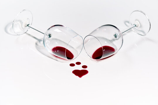 Two high-stemmed glasses with red wine is lying on white background with drops of vine in a shape of heart.
