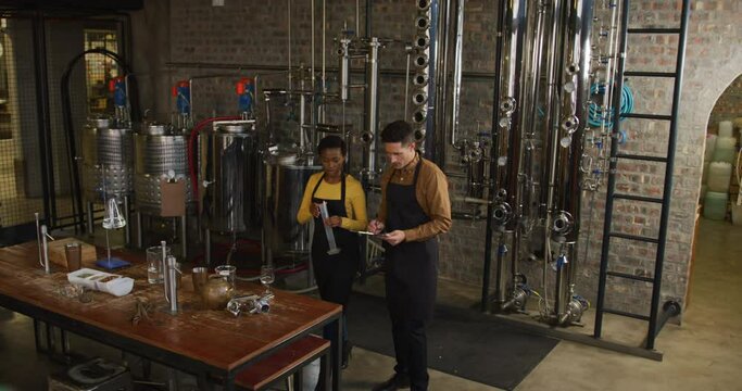 Diverse male and female colleague at gin distillery inspecting equipment, talking and making notes