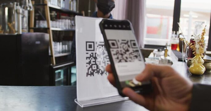 African american woman in face mask serving customer reading qr code with smartphone at cafe bar