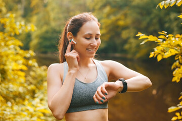 Portrait of cheerful sport young woman outdoors at sunset using earpods and and looking at smartwatch