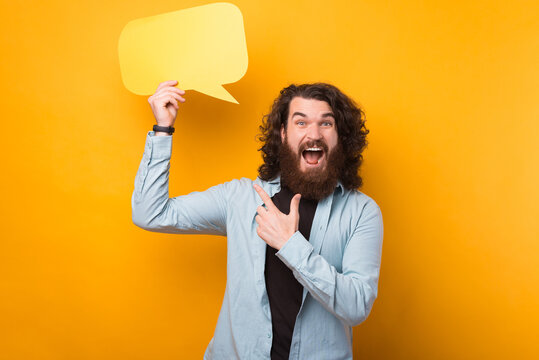 Amazed handsome bearded man with long curly hair pointing at yellow speech bubble