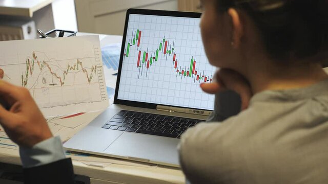Young coworkers comparing sales result graph on paper with chart on computer screen at office. Professional managers examining financial reports during business meeting. Blurred foreground. Rear view