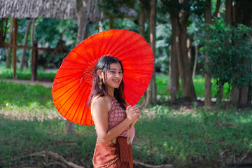 happy Asian woman tourist in Thai traditional dress with red umbrella smiling while looking at beautiful view of nature in green trees forest