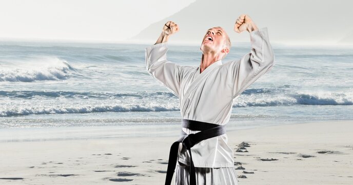 Composition of male martial karate artist with black belt with arms in air on beach with copy space