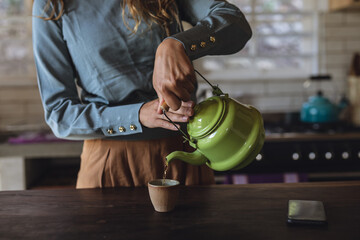 Midsection of caucasian woman standing in cottage kitchen pouring tea from teapot