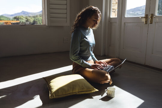 Happy caucasian woman sitting on floor using laptop in sunny cottage living room