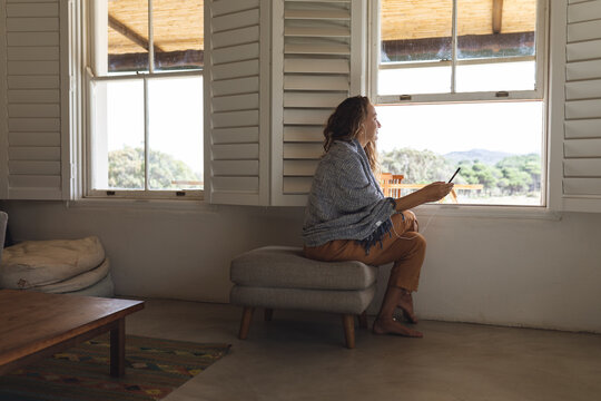 Happy caucasian woman wearing earphones using smartphone sitting by window in cottage living room